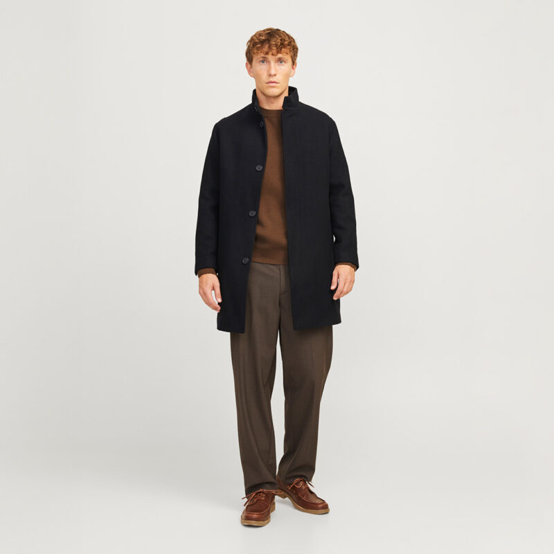 Ein Mann trägt einen schwarzen Mantel, einen braunen Pullover und graue Hosen. Er steht aufrecht vor einem neutralen Hintergrund. Ein Mann trägt einen schwarzen Mantel, einen braunen Pullover und graue Hosen. Er steht aufrecht vor einem neutralen Hintergrund.