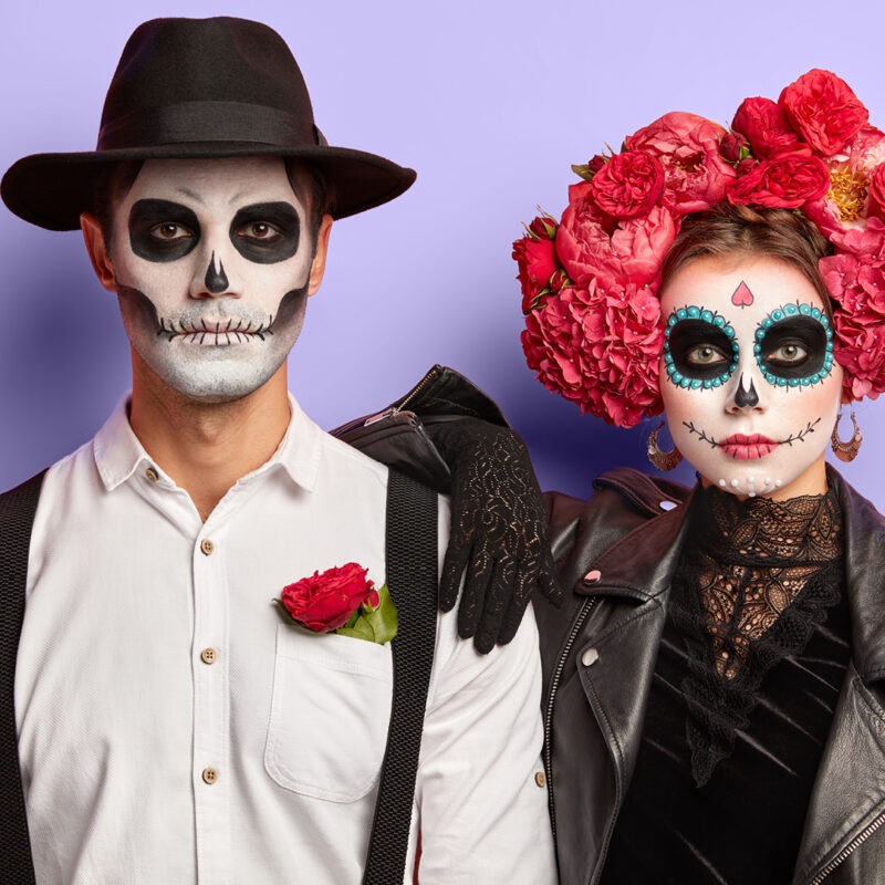 Ein Paar mit Totenschädel-Make-up: ein Mann in weißem Hemd und schwarzem Hut, und eine Frau mit bunten Blumen im Haar und schwarzer Jacke. Ein Paar mit Totenschädel-Make-up: ein Mann in weißem Hemd und schwarzem Hut, und eine Frau mit bunten Blumen im Haar und schwarzer Jacke.