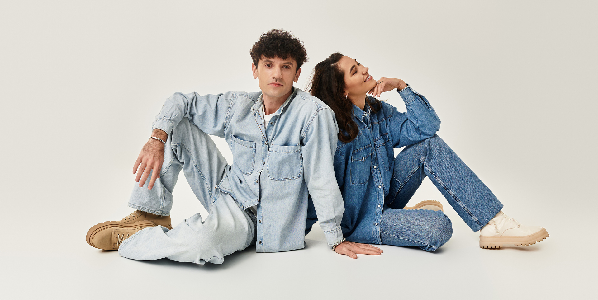 Ein Mann und eine Frau sitzen auf dem Boden, sie tragen Jeanshosen und Jeanshemden sowie beige Boots. Ein Mann und eine Frau sitzen auf dem Boden, sie tragen Jeanshosen und Jeanshemden sowie beige Boots.