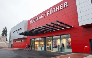 Das Bild zeigt das Gebäude von Modepark Röther mit einer modernen roten Fassade und großen Fenstern. Vor dem Geschäft sind mehrere Personen zu sehen.