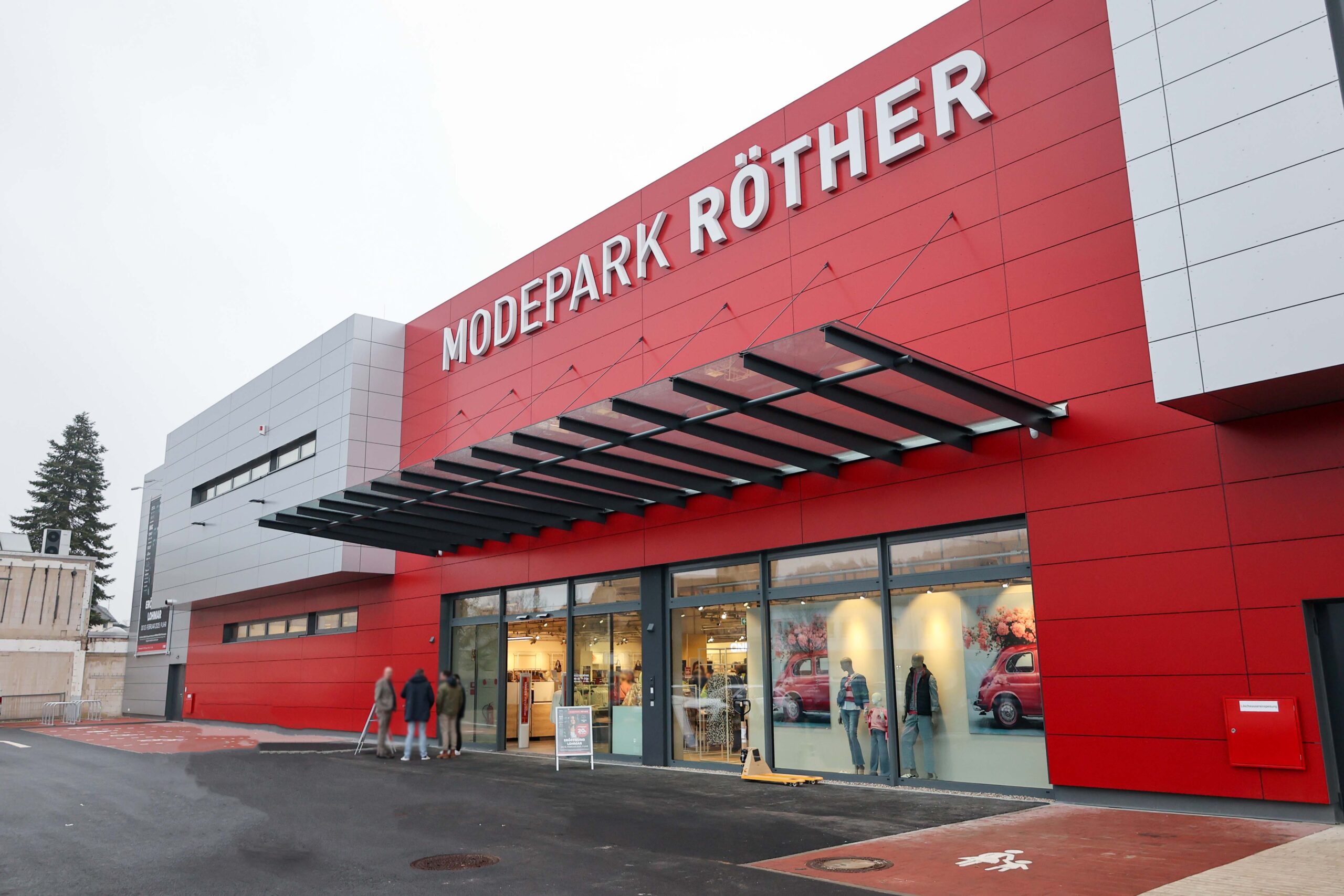 Das Bild zeigt das Gebäude von Modepark Röther mit einer modernen roten Fassade und großen Fenstern. Vor dem Geschäft sind mehrere Personen zu sehen.