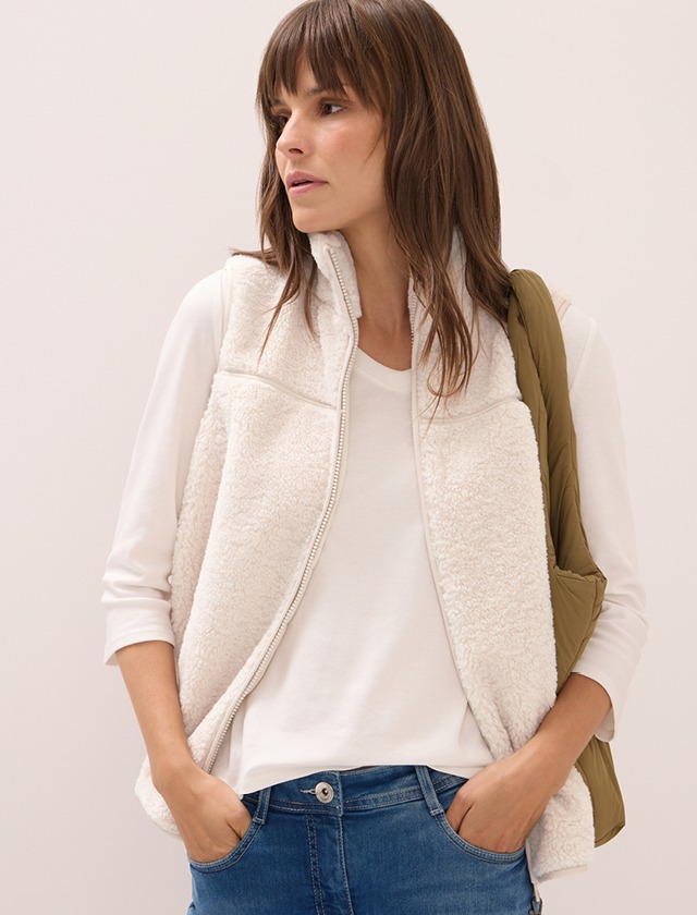 Eine Frau mit langen, braunen Haaren trägt eine helle, pelzartige Weste und ein weißes T-Shirt. Sie hat eine grüne Tasche über der Schulter.