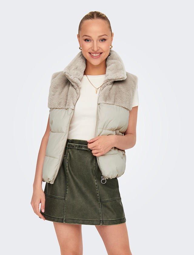 Eine lächelnde Frau trägt eine graue Weste mit Fellkragen und einen grünen Rock.