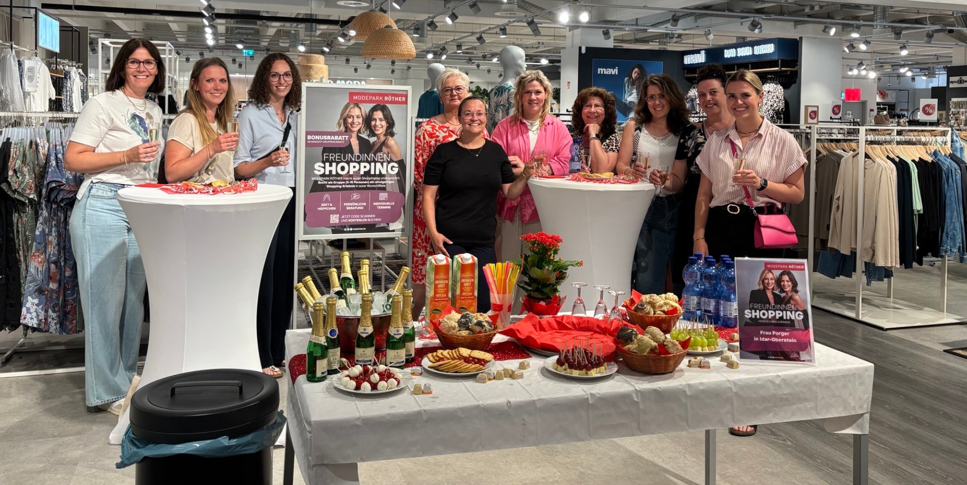 Eine Gruppe von Frauen steht in einem Modegeschäft und feiert mit Sekt. Auf einem Tisch stehen Snacks, Getränke und eine Werbetafel für eine Shopping-Veranstaltung. Eindruck vom Freundinnen-Shopping beim MODEPARK RÖTHER in der Filiale Idar-Oberstein: Persönliche Stilberatung, entspannte Atmosphäre und gemeinsame Shoppingmomente stehen im Mittelpunkt des neuen Eventformats.