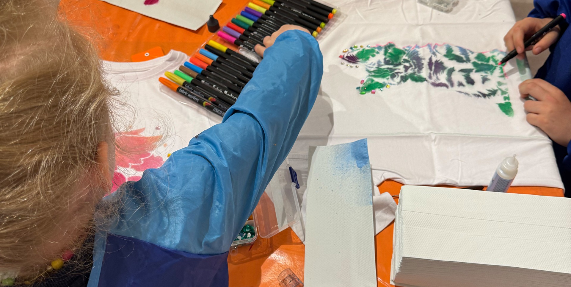Ein Kind malt mit Markern auf einem weißen T-Shirt. In der Nähe liegen weitere Marker und Bastelmaterialien. Ein besonderes Rahmenprogramm während den Kindertagen bei Modepark Röther