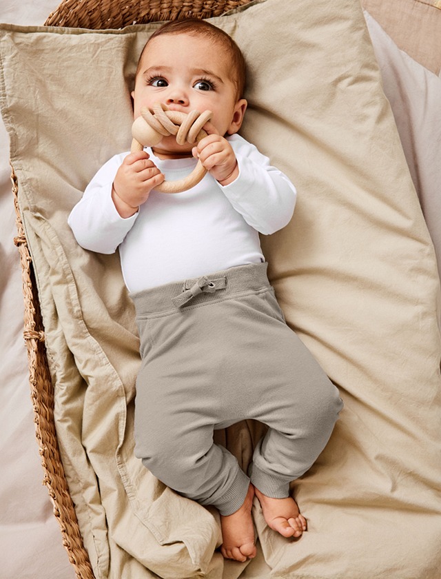 Ein Baby in einem weißen Shirt und grauen Hosen liegt entspannt auf einem Bett. Ein Baby in einem weißen Shirt und grauen Hosen liegt entspannt auf einem Bett.