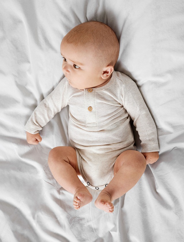 Ein Baby in einem weißen Bodysuit liegt entspannt auf einem Bett. Ein Baby in einem weißen Bodysuit liegt entspannt auf einem Bett.