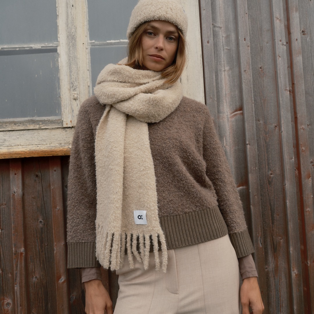 Eine Frau trägt einen braunen pullover, einen beigen Schal und eine bequeme Mütze. Sie steht vor einer Holzfassade und blickt in die Kamera.