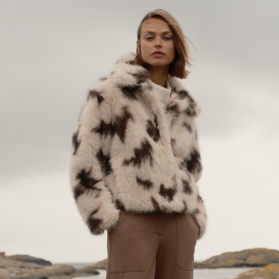 Eine Person trägt einen gefütterten, gemusterten Pelzmantel und steht vor einer bewölkten Himmelshorizont.