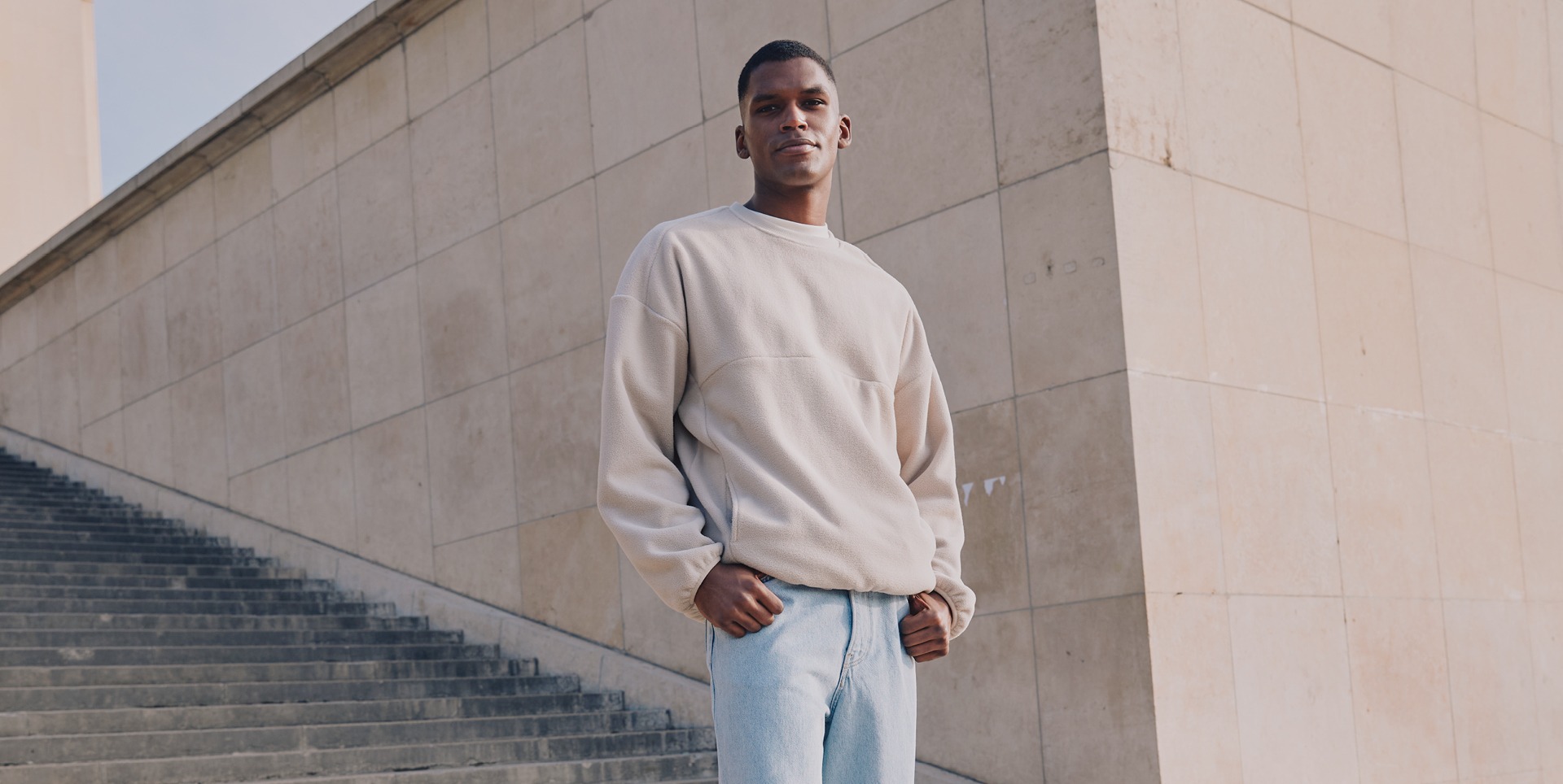 Ein Mann posiert in einem beigen Sweatshirt und blauen Jeans an einer Treppe vor einer Wand.