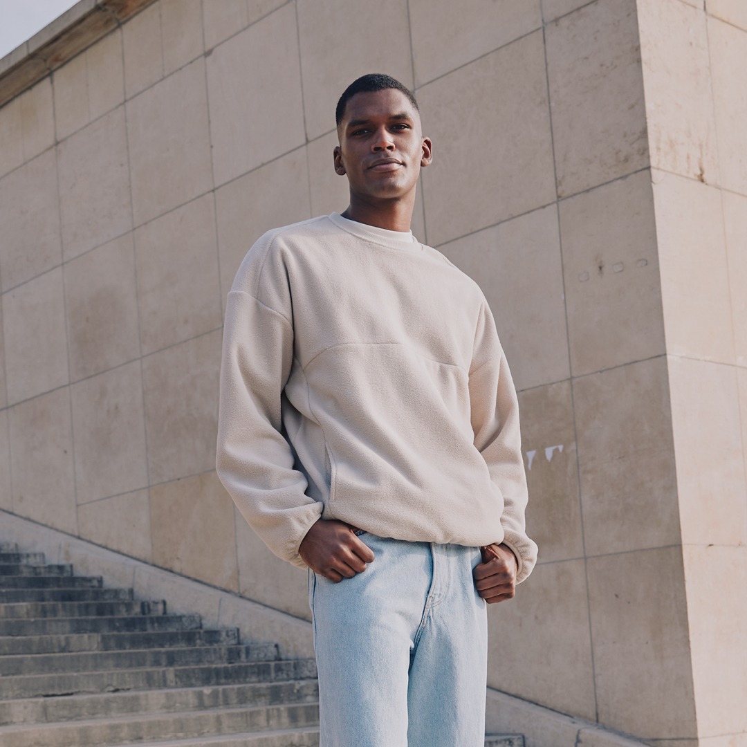 Ein Mann posiert in einem beigen Sweatshirt und blauen Jeans an einer Treppe vor einer Wand.
