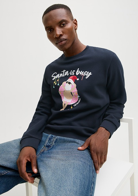 Person sitzt auf weißem Stuhl, trägt dunkelblauen Pullover mit Aufdruck eines Donuts mit Weihnachtsmütze und Schriftzug 'Santa is busy'.