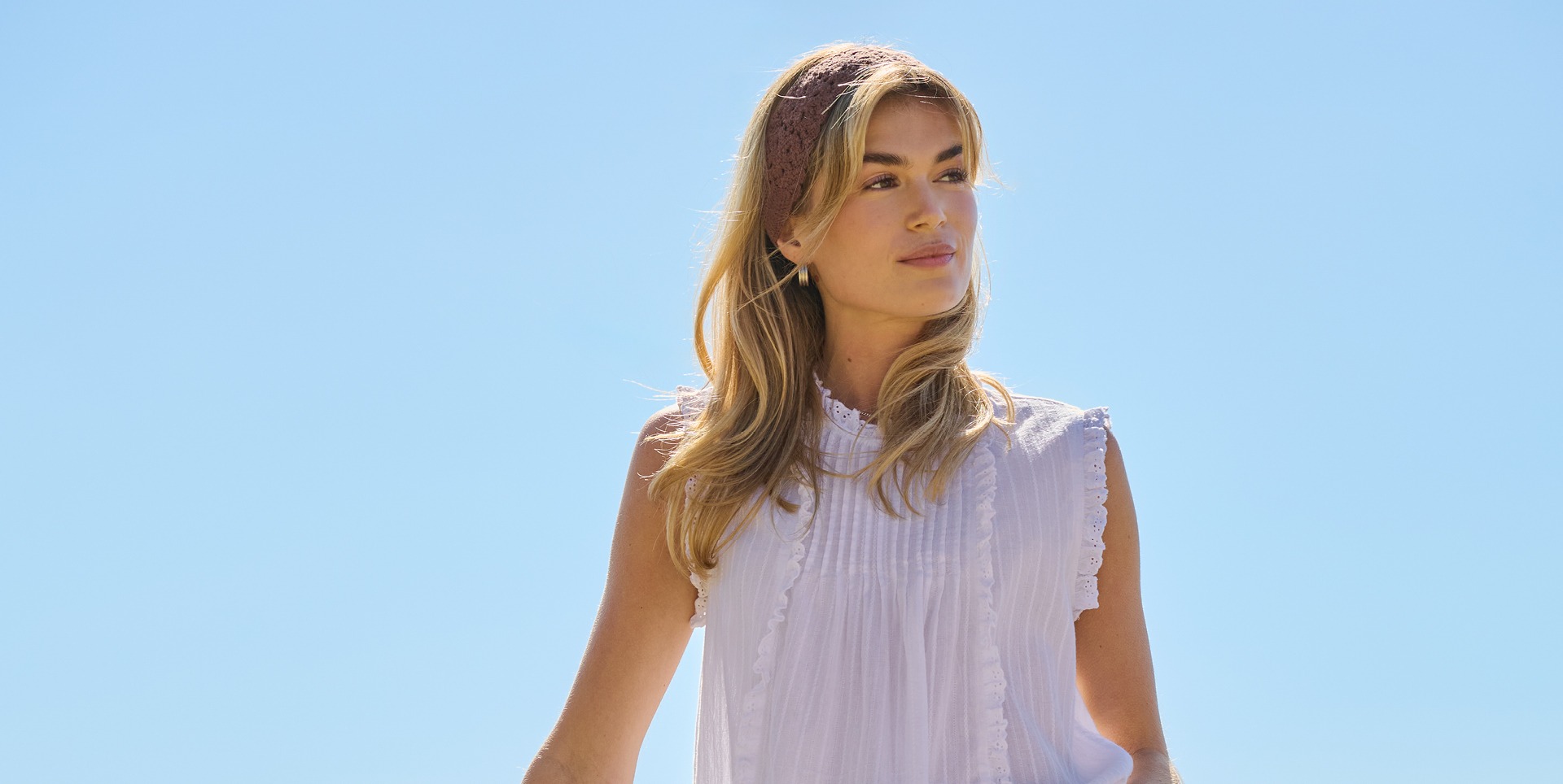 Eine Frau mit langen Haaren trägt ein weißes Oberteil und ein Stirnband, vor einem blauen Himmel. Eine Frau mit langen Haaren trägt ein weißes Oberteil und ein Stirnband, vor einem blauen Himmel.